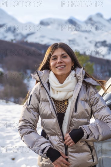 Young woman smiling while adjusting her warm winter jacket with a scarf, standing outdoors against a backdrop of snowy mountains and a clear sky, enjoying a winter trip