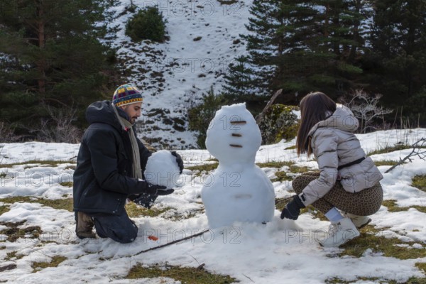 Young couple building a snowman on a sunny mountain slope, laughing and bonding in warm winter clothes amid snowy forest scenery, enjoying playful outdoor holiday recreation