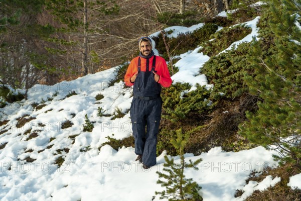 Man in an orange sweater and dark bib pants smiling, enjoying winter nature while walking through a snowy landscape with green bushes and pine trees under sunlight