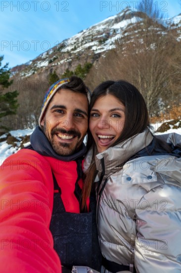 Smiling young man and woman capturing a selfie together during a snowy winter vacation, enjoying the outdoors with a mountain backdrop under a clear blue sky