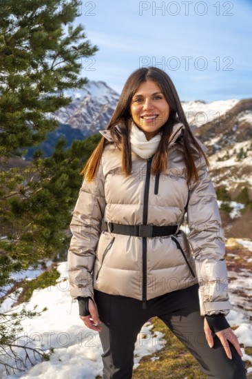 Young woman smiling at camera in snowy mountain landscape, wearing warm jacket and enjoying a winter travel adventure amid pine covered peaks and clear blue sky