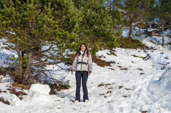 Young woman wearing warm winter clothes and a shiny jacket smiling while standing on a snowy mountain path with pine trees in a natural outdoor landscape under blue sky