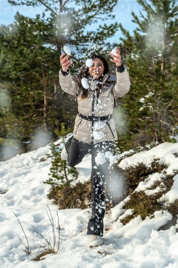 Young woman in casual winter clothes laughing and throwing snowballs in a sunlit snowy forest, enjoying outdoor activity, carefree fun and healthy seasonal adventure