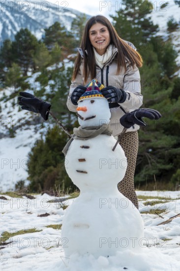 Young woman smiling while creating a snowman with a hat, scarf, carrot nose, and tree branch arms, celebrating winter vacations outdoors in a snowy mountain landscape