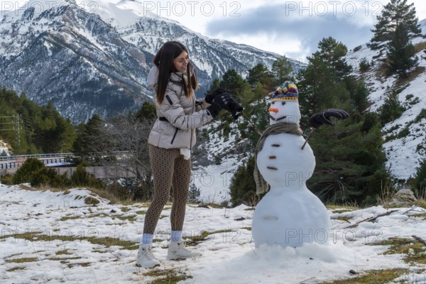 Young woman actively building a snowman outdoors with snow capped mountains and pine trees in the background, enjoying a fun winter leisure activity during cold weather
