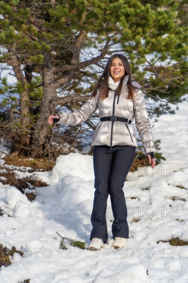 Young woman smiling and spreading her arms, dressed in warm winter clothing with a metallic jacket and black pants, walking through fresh snow on a sunny day near pine trees