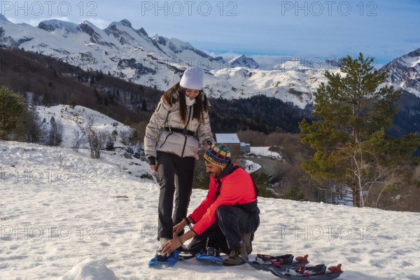 Couple preparing their snowshoes for a winter hike, strapping them onto their boots with snowy mountains and trees in the background, enjoying an outdoor adventure