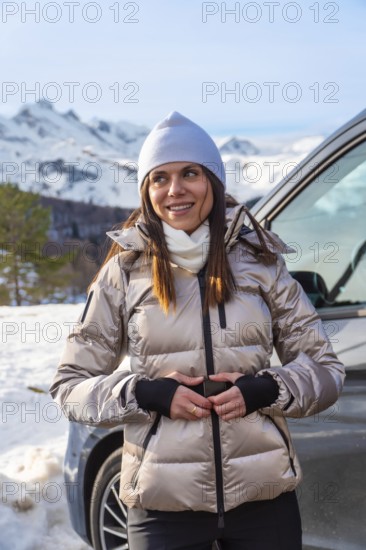 Woman wearing a puffer jacket and beanie smiling while standing by a camper van, enjoying the scenic snowy mountains and clear blue sky during a winter road trip or vacation