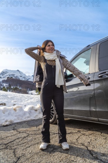 Young woman standing on a snowy mountain road next to a dark vehicle, wearing winter clothes including a leopard print top and black jumpsuit, looking excited during a road trip adventure