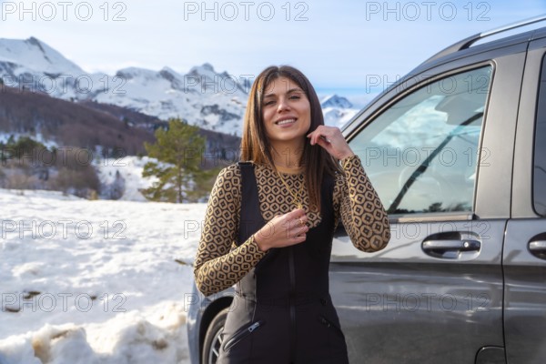 Young woman smiling while standing next to a car, enjoying a winter road trip adventure with snow covered mountains and trees in the background, experiencing nature