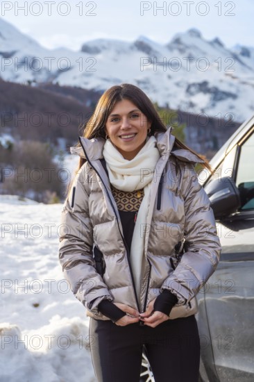 Young woman smiling while zipping up her warm jacket, standing outdoors in a snowy mountain landscape, preparing for winter adventure and travel during bright daylight