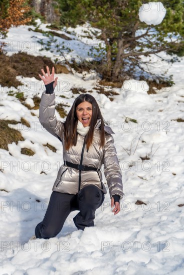 Young woman kneeling in snow with a playful expression, throwing a snowball against a backdrop of snow covered terrain and trees, embodying winter fun and outdoor activity