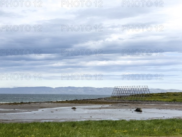 Coast with green shore, wooden racks for drying stockfish and cloudy sky over the sea, Nesseby, Varangerbotn, Finnmark, Norway