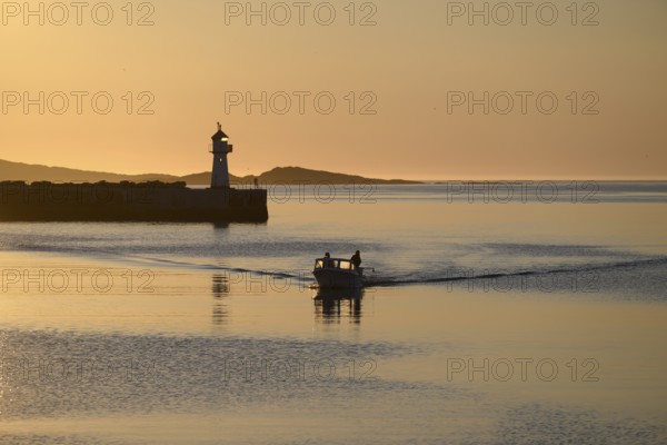 A small motorboat sails through the harbor entrance with lighthouse and midnight sun, Vardø, Finnmark, Norway