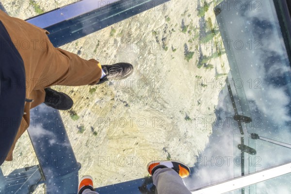 Tourists are enjoying a unique perspective, walking on a glass floor high above a rocky mountain valley, offering a breathtaking view of the landscape below