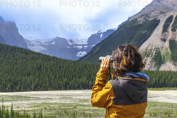 Female hiker wearing a yellow jacket using binoculars while observing a glacier in the canadian rockies along the icefields parkway, surrounded by lush pine forest and mountains