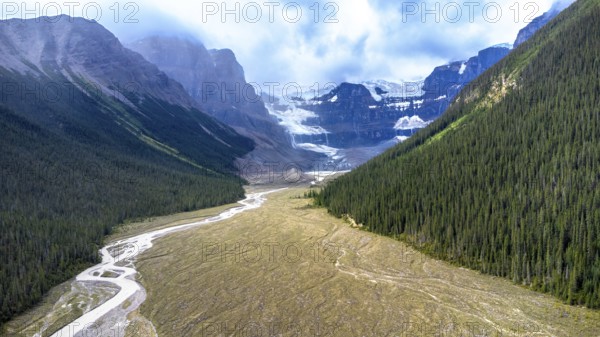 Sun shining on athabasca glacier in jasper national park, alberta, canada, with conifer forest and meltwater river flowing through valley under cloudy sky