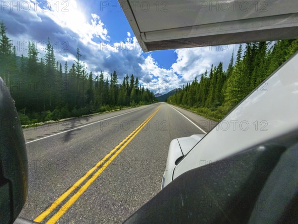 White vehicle driving along icefields parkway, surrounded by lush forests, under a bright blue sky with white clouds, offering a breathtaking view of the canadian rockies