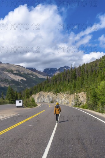 Hiker walking on the icefields parkway in the canadian rockies with a camper van parked on the side of the road and mountains and forest in the background under a blue sky with clouds