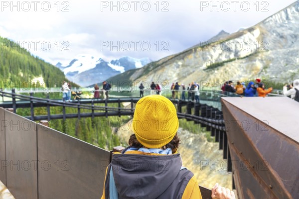 Tourist wearing a yellow beanie stands on a viewing platform, admiring the stunning view of athabasca glacier and the surrounding mountains in jasper national park