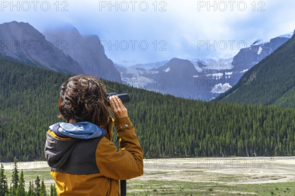 Tourist wearing an orange jacket uses binoculars to observe a glacier in the canadian rockies, surrounded by a coniferous forest and mountains