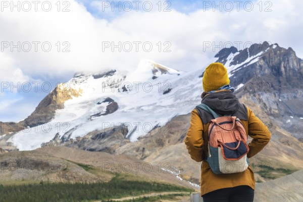 Female tourist wearing a yellow woolen hat and carrying a backpack is contemplating athabasca glacier in jasper national park, alberta, canada, surrounded by stunning natural beauty