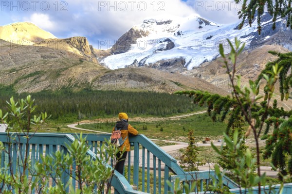 Tourist wearing a yellow beanie and jacket, leaning against a wooden fence, admiring the breathtaking athabasca glacier in jasper national park, alberta, canada