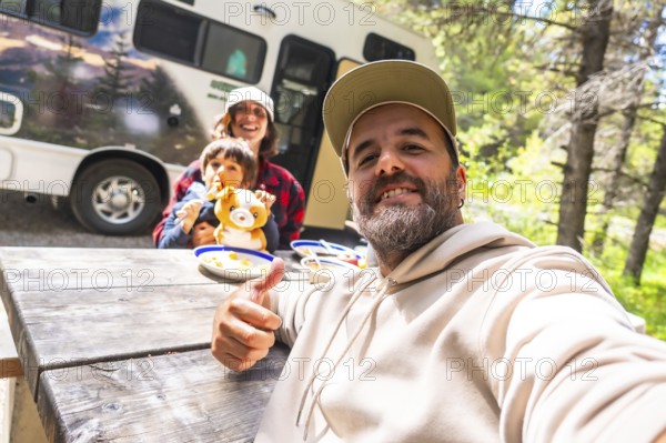Happy family taking a selfie while having lunch on a wooden table near their camper van in the woods, enjoying their vacation in banff national park, canadian rockies