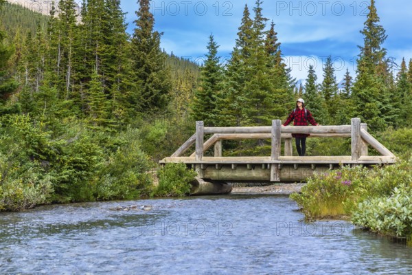 Female hiker enjoying the view from a wooden bridge over a river flowing from bow lake in banff national park, canadian rockies, alberta, canada, on a sunny day with blue sky and clouds