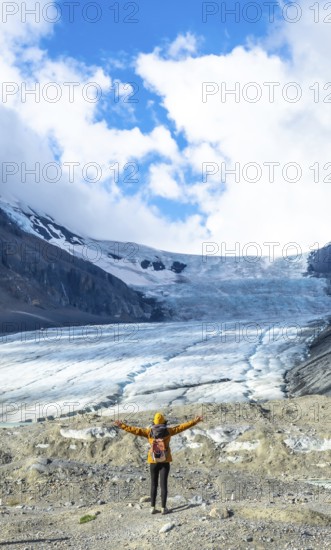Female hiker with backpack and yellow jacket is enjoying the breathtaking view of the athabasca glacier in jasper national park, canadian rockies, alberta, under a beautiful cloudy sky