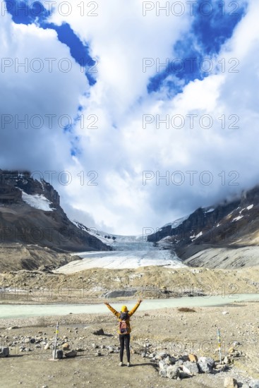 Female hiker with backpack and yellow jacket raising arms in front of the athabasca glacier, in the canadian rockies, jasper national park, alberta, canada, celebrating the beauty of nature