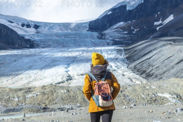 Female tourist in a yellow beanie and jacket, carrying a backpack, admiring the melting athabasca glacier in jasper national park, alberta, highlighting climate change