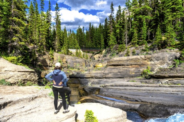 Two tourists admiring the stunning view of mistaya canyon in banff national park, featuring unique rock formations, lush forest, and a bridge on a sunny day