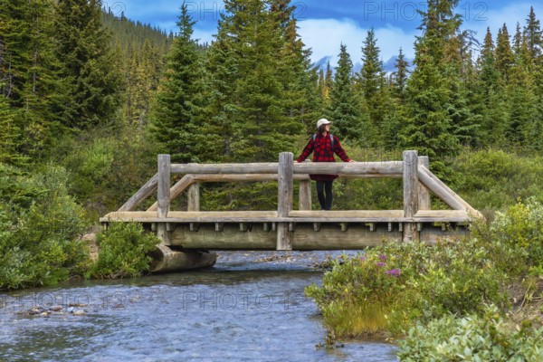 Female hiker wearing red plaid shirt and white hat is leaning on a wooden bridge railing over a clear flowing stream, enjoying the scenic view of bow lake in banff national park, alberta, canada