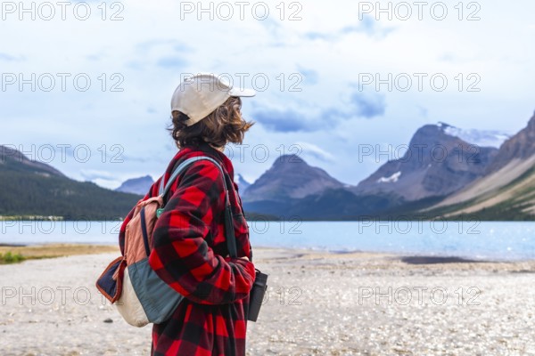 Young hiker standing on the shore of turquoise bow lake, contemplating the majestic mountains of banff national park in the canadian rockies, on a cloudy summer day