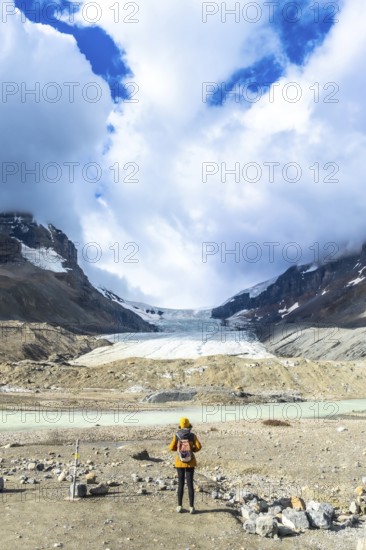 Female tourist wearing yellow jacket and backpack admiring the stunning view of the athabasca glacier in the canadian rockies, jasper national park, alberta, under a cloudy blue sky