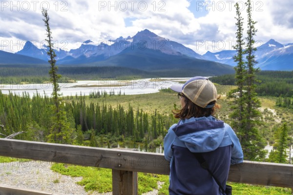 Tourist wearing a baseball cap and light blue jacket leaning on a wooden railing, taking in the panoramic view of the north saskatchewan river and canadian rockies in banff national park