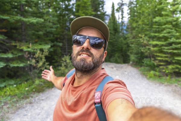 Bearded tourist with sunglasses and cap taking a selfie with arms outstretched at mistaya canyon in banff national park, surrounded by lush pine forest and a gravel path