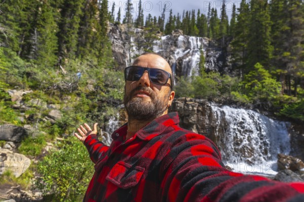 Tourist wearing sunglasses and red and black shirt taking selfie with outstretched arm in front of tangle creek falls in jasper national park, alberta, canada, during sunny summer day