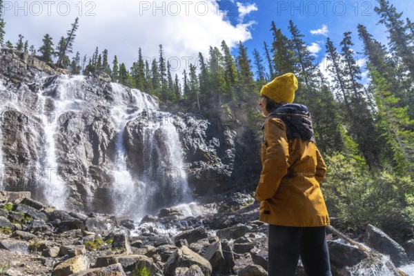 Female hiker wearing yellow beanie and jacket is admiring cascading waters of tangle creek falls in jasper national park, canadian rockies, alberta, canada, during sunny summer day