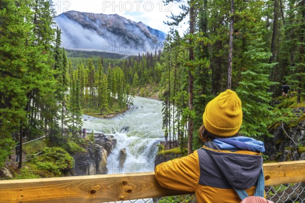 Young hiker wearing a yellow beanie and jacket admiring the stunning view of sunwapta falls in jasper national park, surrounded by the majestic canadian rockies