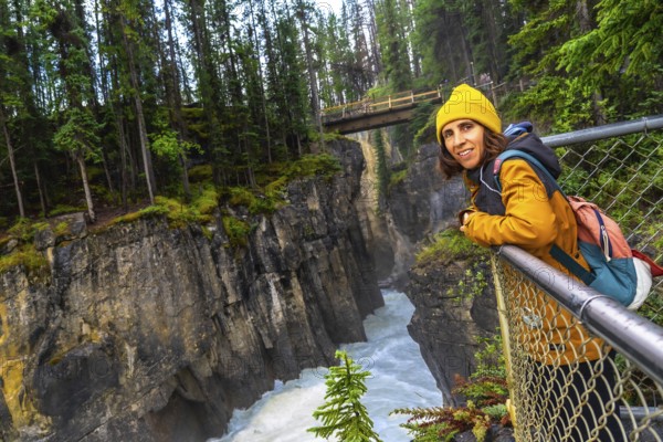 Female tourist in a yellow jacket and beanie leaning on a metal railing, admiring the stunning view of sunwapta falls in jasper national park's lush landscape