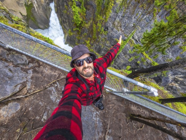 Photographer wearing sunglasses and a bucket hat takes a selfie at sunwapta falls in jasper national park, extending one arm for a wider view of the waterfall and surrounding forest