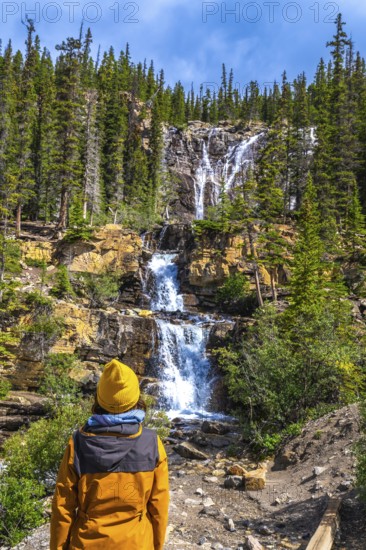 Hiker in a yellow jacket and beanie contemplating the cascading waters of tangle creek falls in jasper national park, surrounded by the stunning canadian rockies