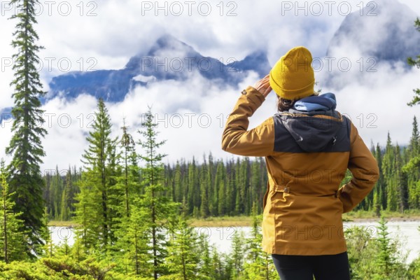 Hiker shielding her eyes while admiring the breathtaking vista of the canadian rockies along the icefields parkway, surrounded by lush pine trees and a flowing river