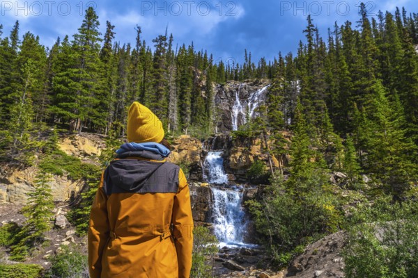Female hiker in a yellow beanie and jacket enjoying breathtaking views of tangle creek falls cascading down rocky slopes amid lush pine forests in jasper national park