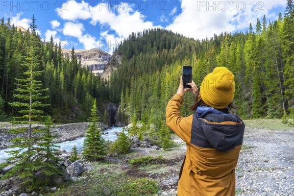 Hiker capturing smartphone images of a turquoise waterfall flowing through a lush pine forest along the scenic icefields parkway in banff national park, alberta