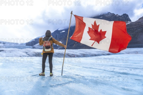 Female tourist is holding large canadian flag while standing on surface of athabasca glacier in jasper national park, alberta, canada, celebrating iconic natural landmark