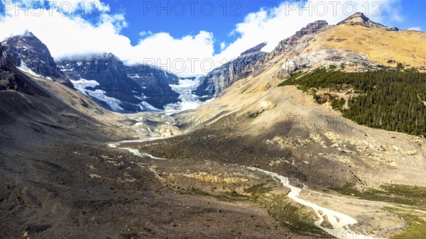 Sun shining on athabasca glacier in the canadian rockies with clouds and blue sky reflecting on melting glacial ice feeding a river with conifer forest, rocks, and mountain peaks