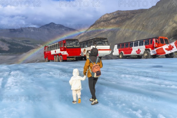 Tourists walking on the surface of athabasca glacier in jasper national park, alberta, canada, with large red tour buses parked nearby and a vibrant rainbow arching across the sky